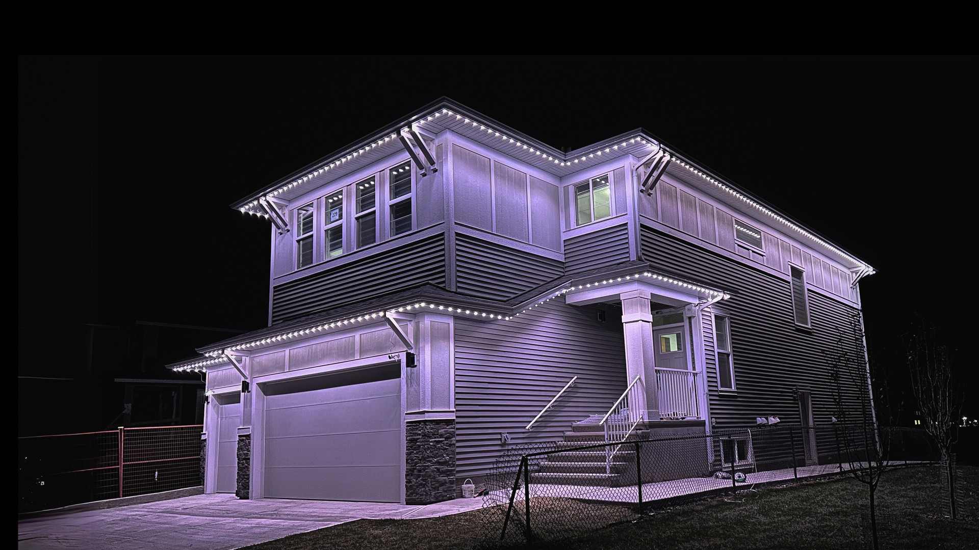 A grey two-storey home at night lit in crisp cool white, the light following every roofline and the covered entry.