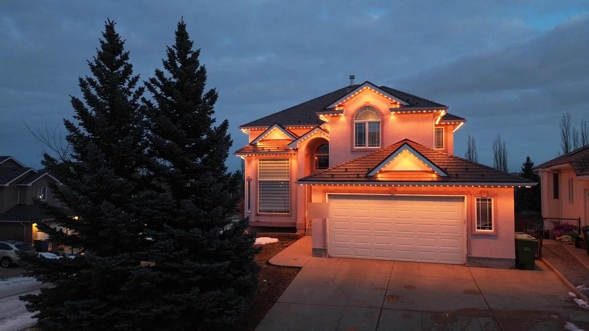 A two-storey home at dusk under a blue Alberta sky, rooflines and garage picked out in warm amber light, a tall spruce in the foreground.
