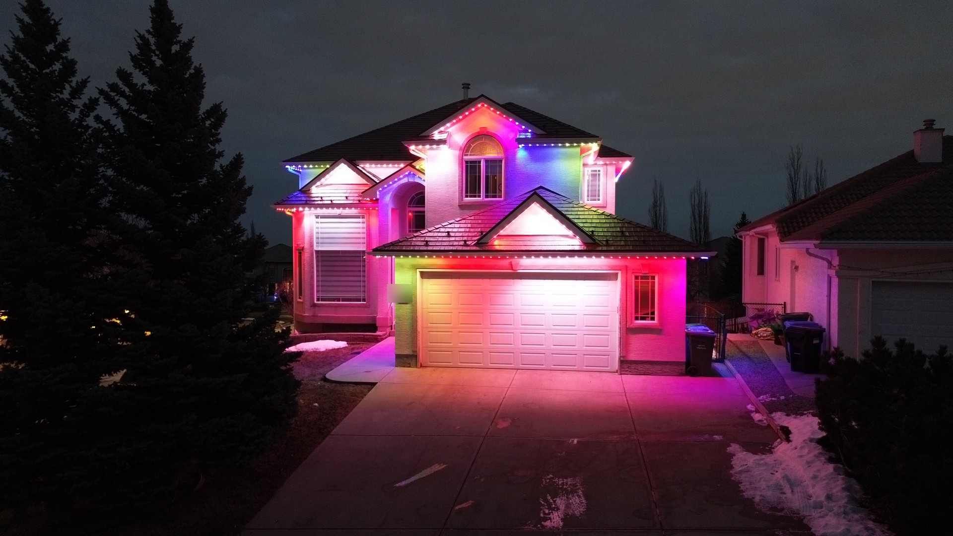 The same home after dark in a bright multicolour scene, colour reflecting off snow on the driveway.