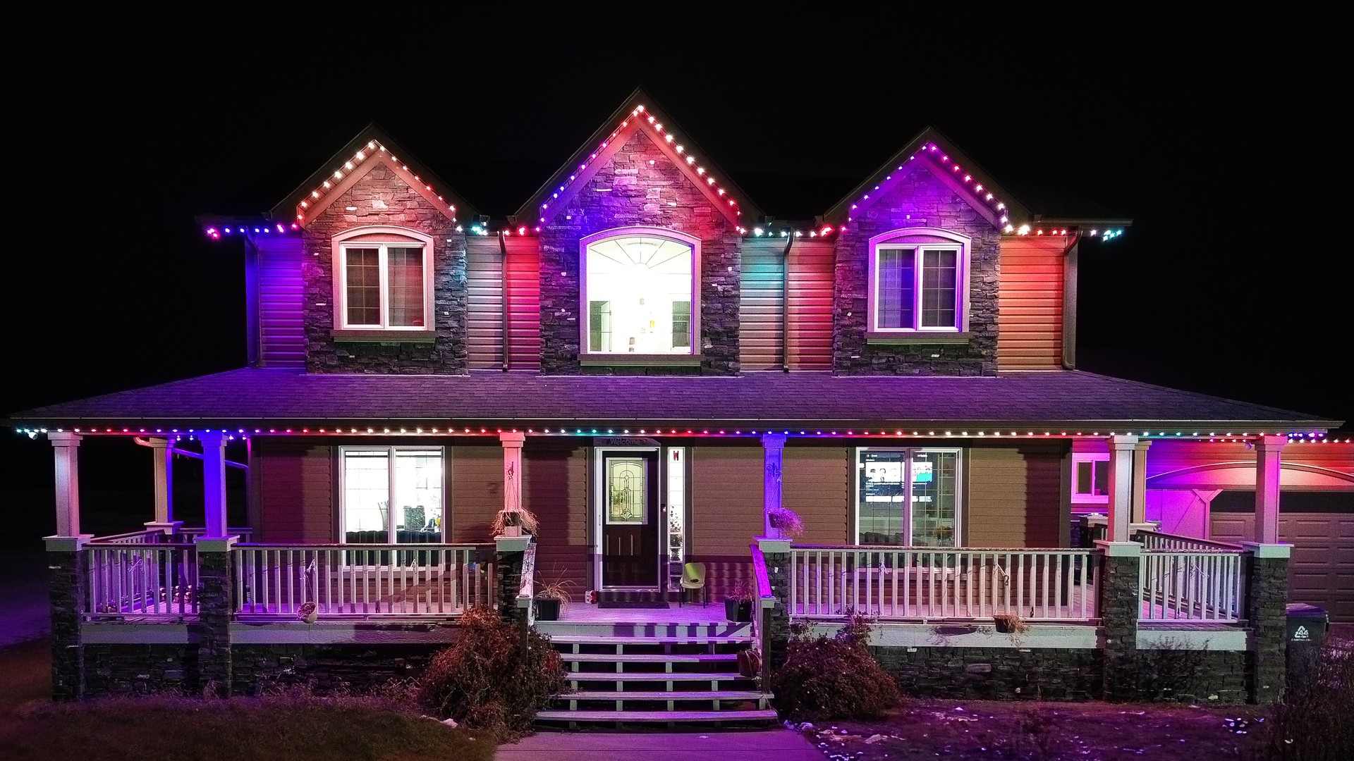 A two-storey home with a wraparound porch lit in shifting purple, pink and green, the colour spilling onto the porch rails and front lawn.