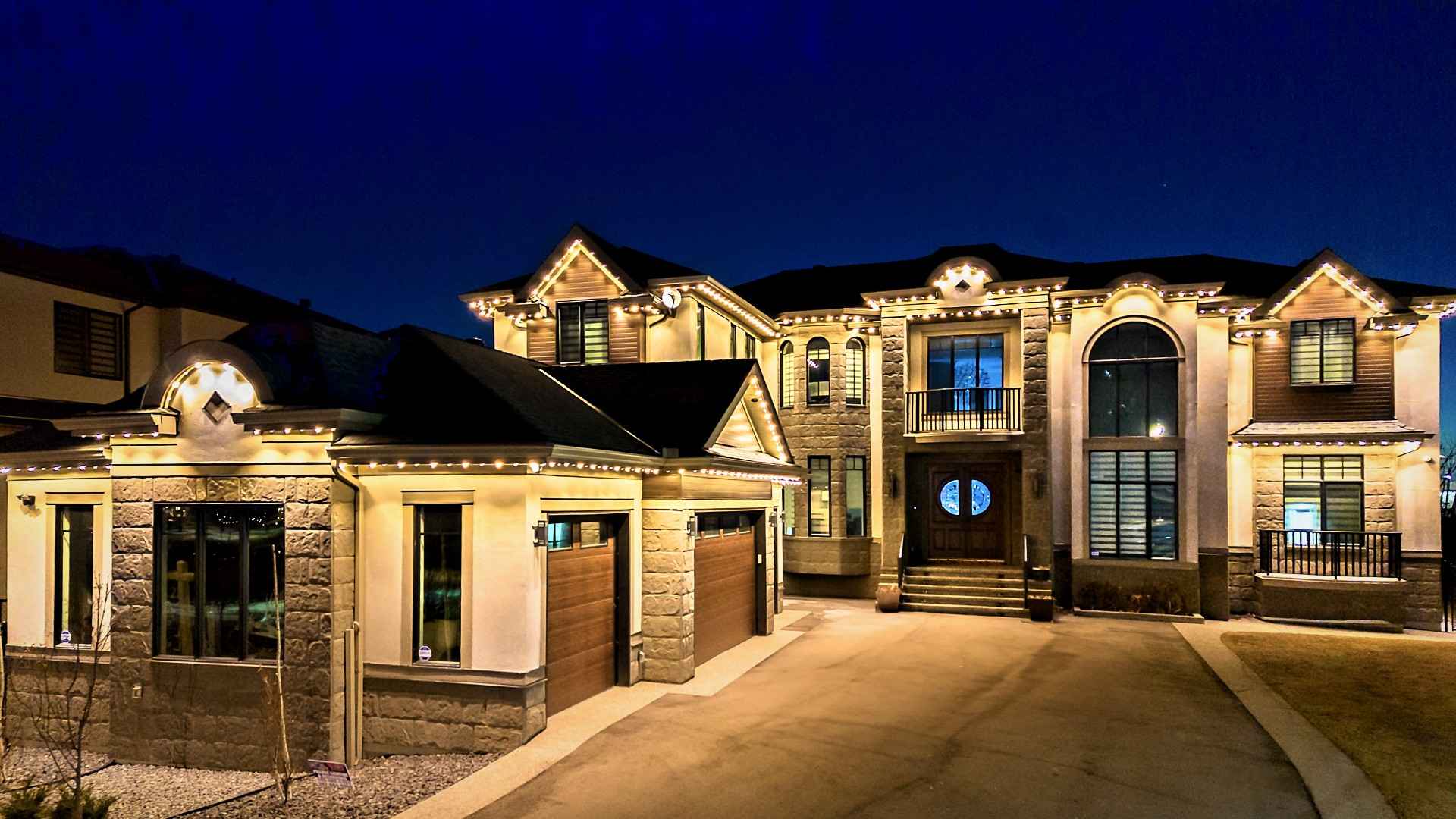 A large stone-and-stucco home at blue hour, every gable, arch and eave outlined in even warm-white light, the driveway sweeping up to a lit front entrance.