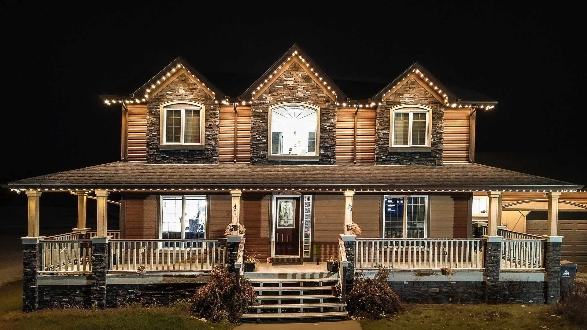 A two-storey Alberta home at night with three dormer gables and a wraparound porch, every roofline traced by a run of individually controlled lights.
