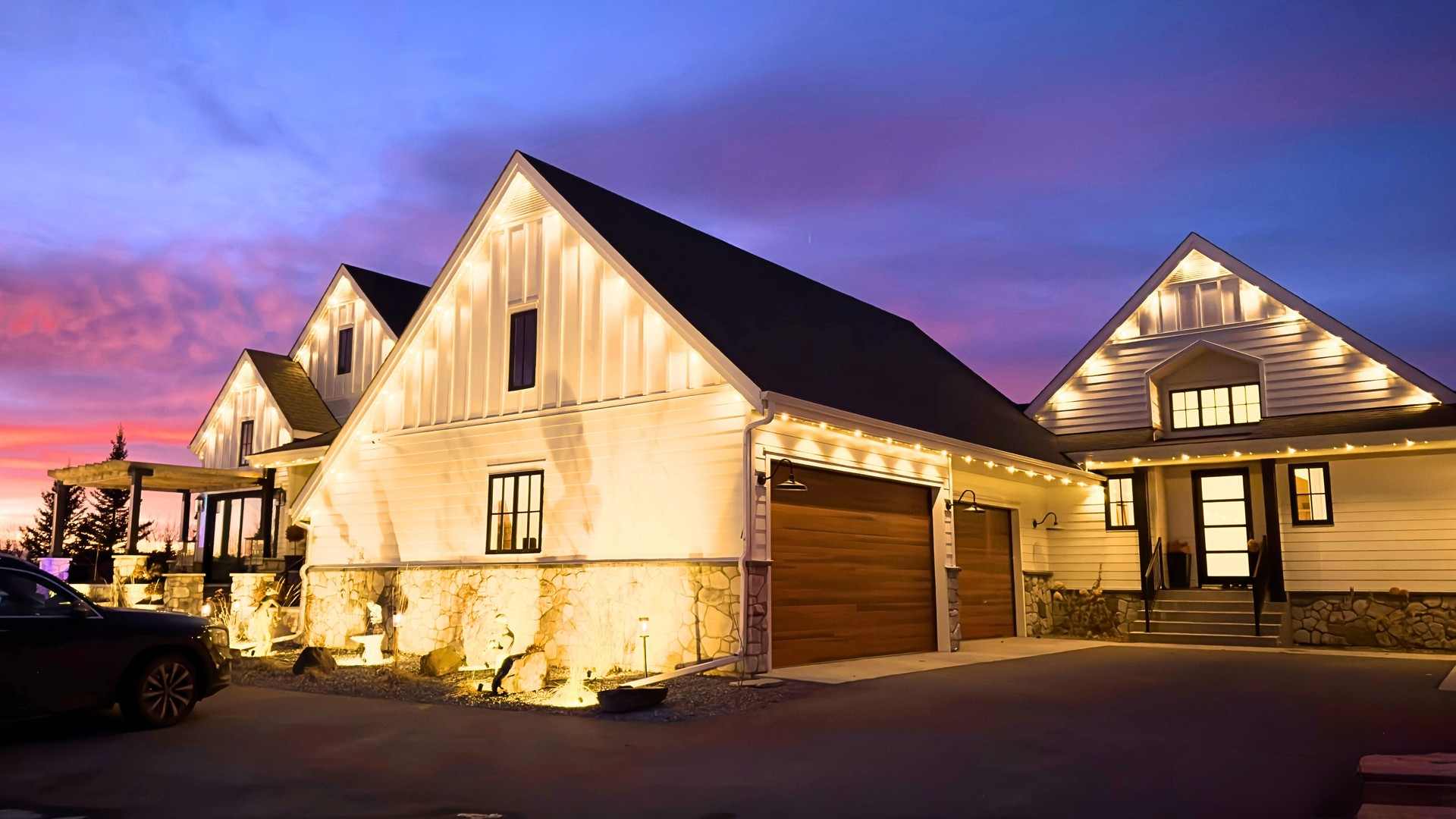 A white modern farmhouse at sunset, its steep gables and garage lit in warm white against a pink and violet Alberta sky. In the remaining daylight the track itself is barely visible along the roofline.